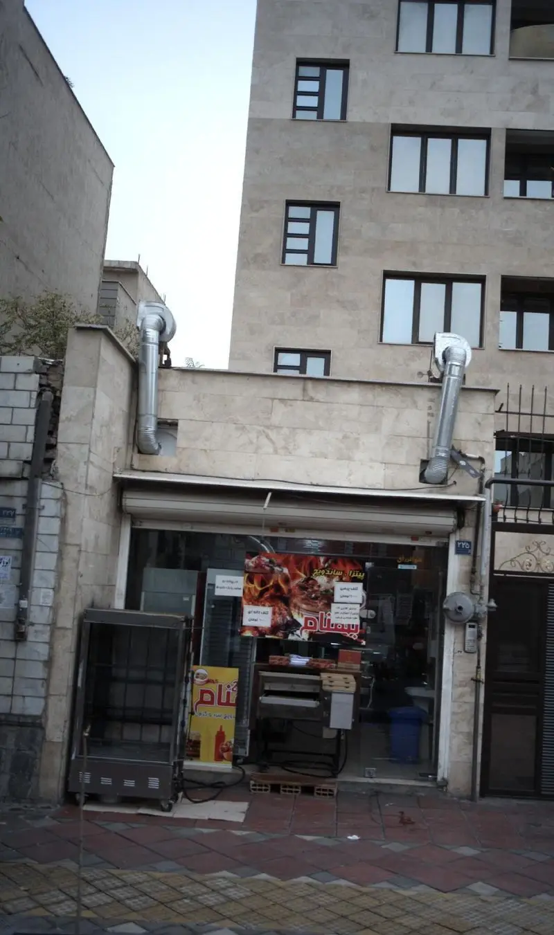 نمای بیرونی فست فود بهنام، یک رستوران پیتزا و ساندویچ در شهر، با تابلوهای جذاب غذا، دودکش‌های فلزی و ویترین شیشه‌ای.