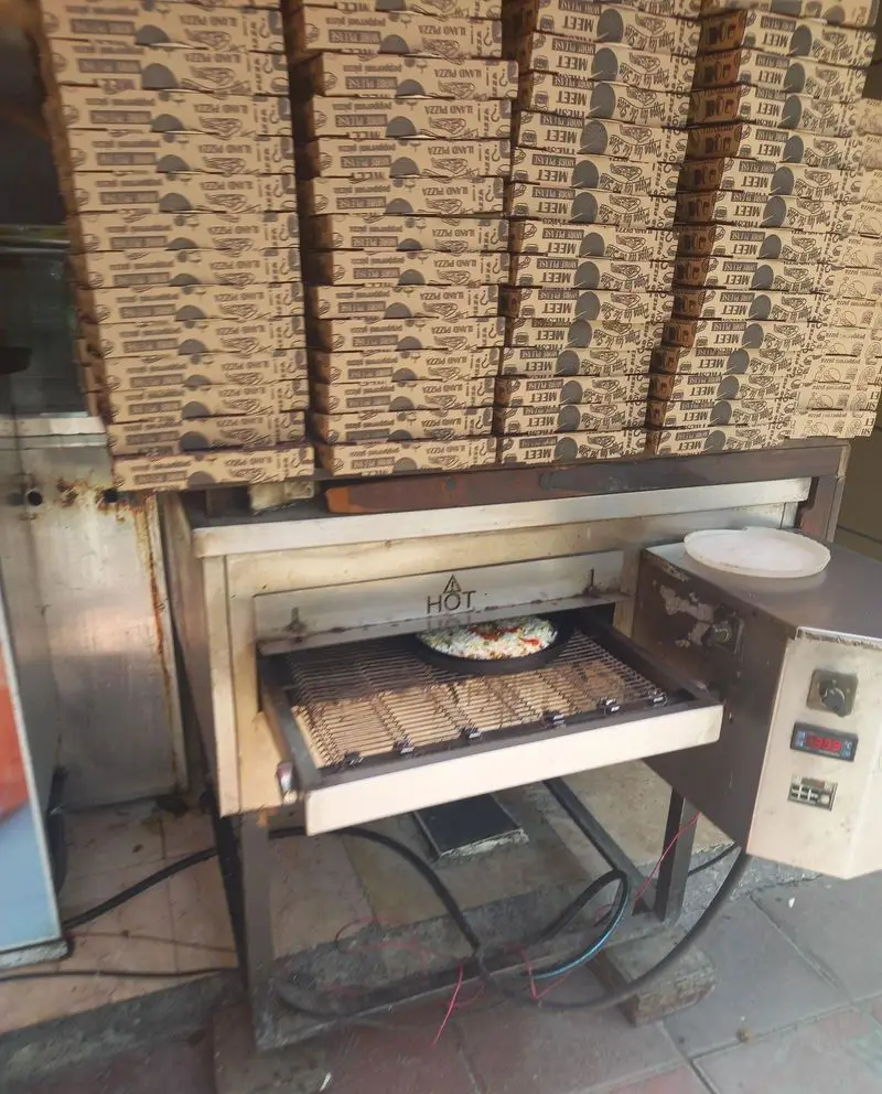 تصویری از فر پیتزاپزی فست فود بهنام در حال پخت پیتزای داغ، با انبوهی از جعبه‌های پیتزا لند و میت فرش آماده برای بسته‌بندی.