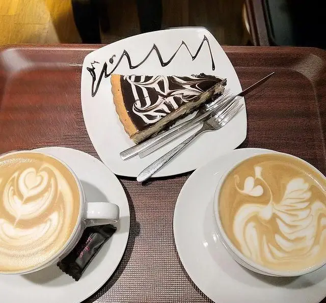 تصویری جذاب از دو فنجان قهوه لاته آرت (قلب و قو) و کیک شکلاتی در کافه رستوران لی وان، انتخابی عالی برای دسر و نوشیدنی‌های گرم.