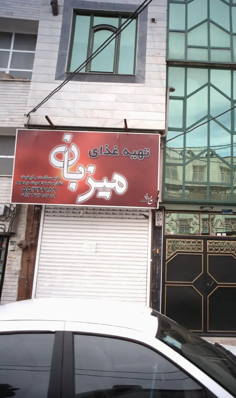 نمای بیرونی تهیه غذای میزبان: تابلوی قرمز با نام کسب و کار، شعار سلامت و کیفیت غذا و شماره تماس، کرکره مغازه بسته است.