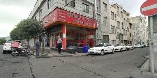 مرغ سوخاری و پیتزا پاپینوس: نمای بیرونی مغازه فست فود در یک روز ابری، با موتورسیکلت تحویل غذا و مشتریان در کنار خیابان.