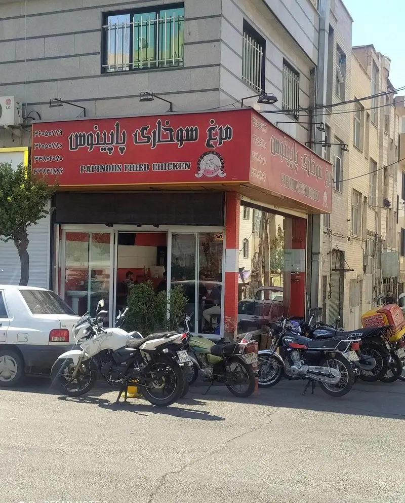 نمایی از رستوران فست فود پاپینوس متخصص مرغ سوخاری، با تابلوی بزرگ و قرمز رنگش، خودرو و موتور سیکلت های متعدد پارک شده در خیابان.