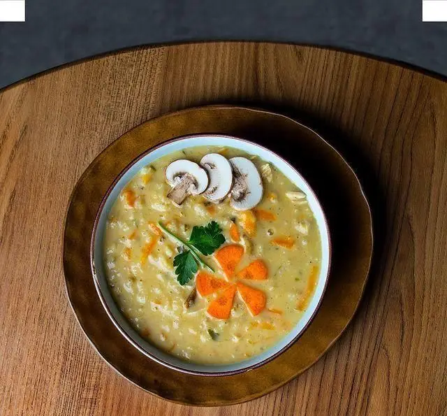 سوپ خامه ای قارچ و مرغ خوشمزه کافه رستوران هفت آسمان، با تزیین هنرمندانه هویج و قارچ تازه، آماده سرو روی میز چوبی.
