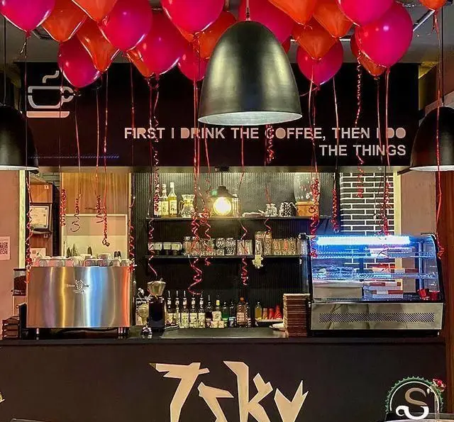 کافه رستوران هفت آسمان: فضای داخلی با دستگاه اسپرسو، دسرهای متنوع و بالون‌های رنگی. شعار جذاب قهوه روی دیوار.