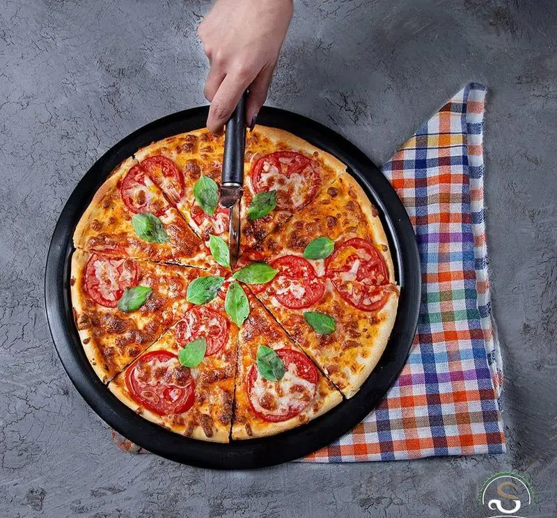 برش زدن پیتزا مارگاریتا داغ و خوشرنگ با پنیر، گوجه تازه و برگ‌های معطر ریحان، مخصوص کافه رستوران هفت آسمان.