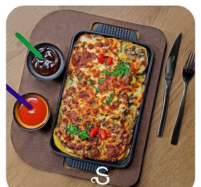 غذای پنیری و گراتن خوشمزه کافه رستوران هفت آسمان، سرو شده در ظرف مستطیلی با سس‌های متنوع روی سینی چوبی و کارد و چنگال.