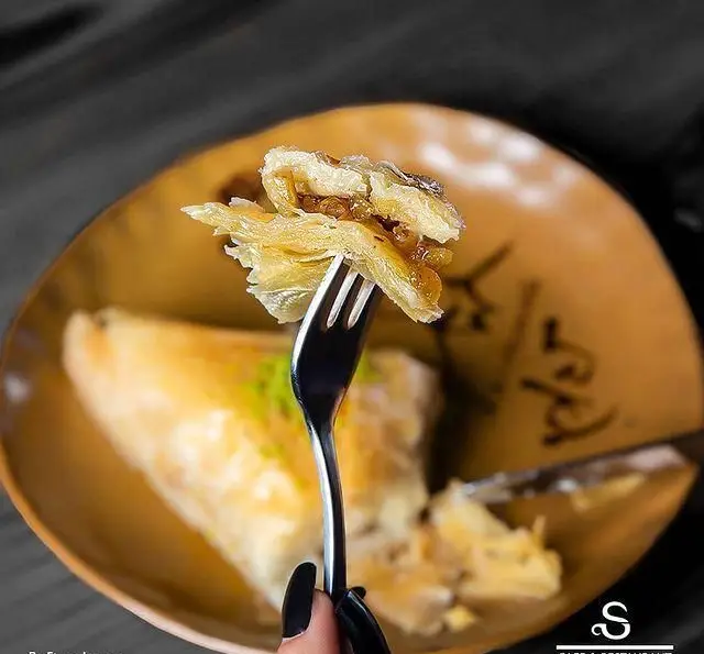 تکه‌ای دسر لایه‌ای خوشمزه (شبیه باقلوا) روی چنگال، نمادی از شیرینی‌های بی‌نظیر کافه رستوران هفت آسمان.