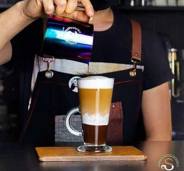 باریستای کافه رستوران هفت آسمان در حال ریختن قهوه لایه‌ای با پیچر رنگین؛ ترکیبی هنرمندانه از اسپرسو و شیر.