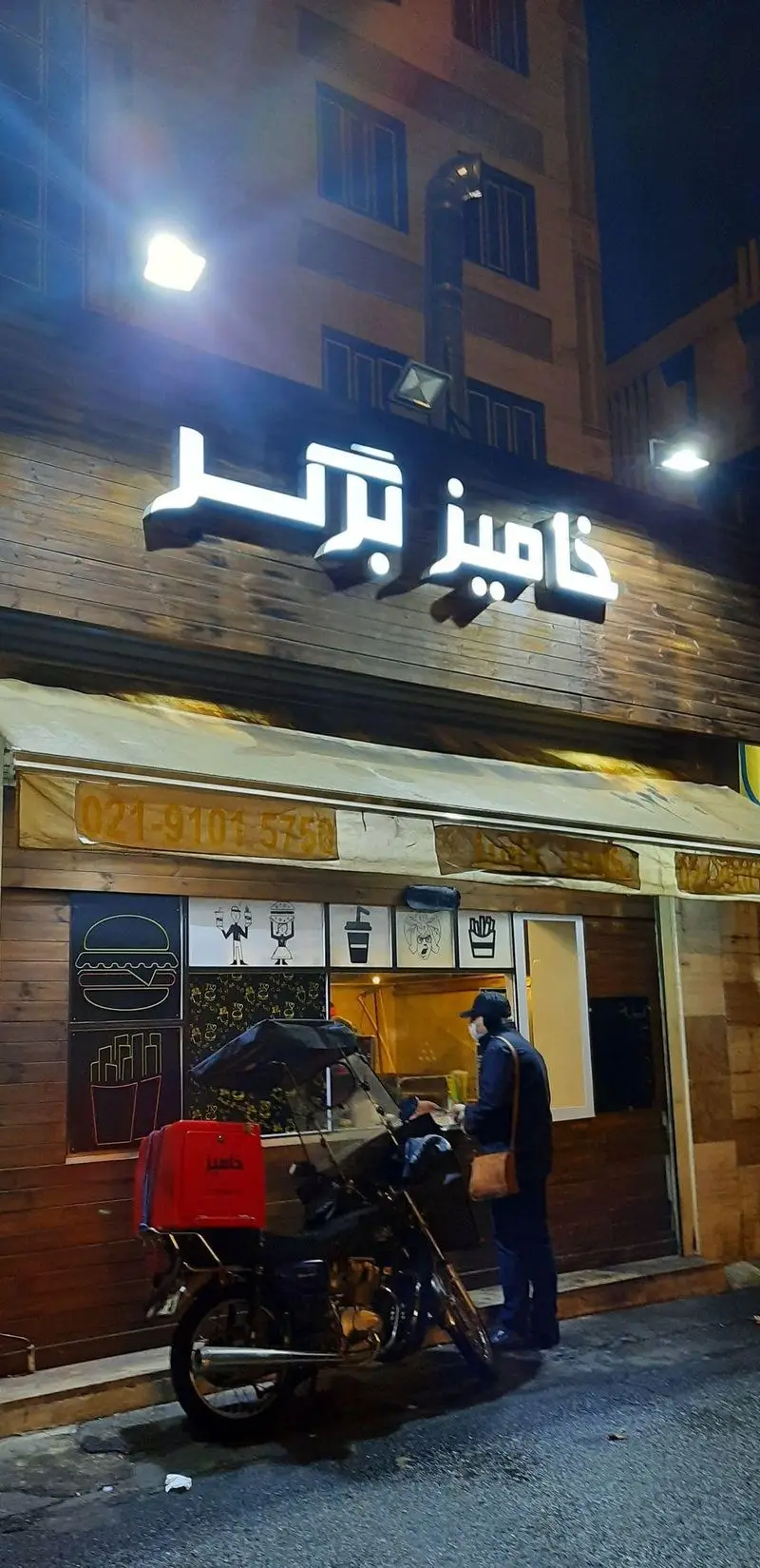 تصویری از نمای شبانه شعبه خامیز برگر در تهران، با نورپردازی جذاب و موتورسیکلت دلیوری. بهترین فست فود و همبرگر را آنلاین سفارش دهید.