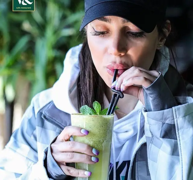 زن جوان در کافه رستوران ویند از نوشیدن اسموتی سبز و خنک با برگ نعناع لذت می‌برد. منوی متنوع کافه و محیط دلنشین.