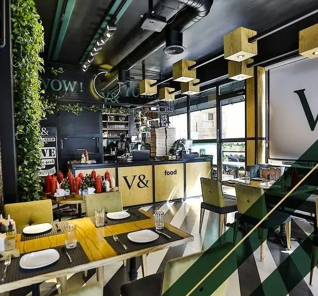 تصویر فضای داخلی شیک و مدرن کافه رستوران ویند. دکوراسیون منحصر به فرد با میز و صندلی‌های راحت، پیشخوان "V& food"، و گیاهان آویزان.