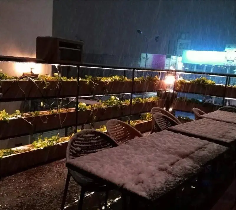 فضای باز کافه رستوران ویند در شبی برفی و زمستانی، با میز و صندلی‌های پوشیده از برف، نورپردازی گرم و گلدان‌های سبز. محیطی دنج و تماشایی برای مشتریان.