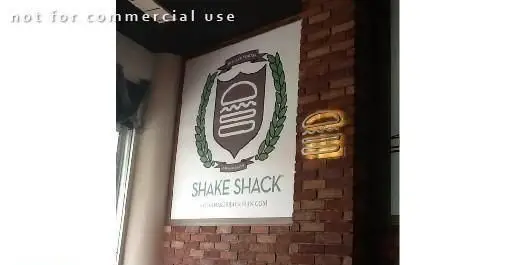 تابلو رستوران فست فود Shake Shack با لوگوی همبرگر در سپر و شاخه زیتون، روی دیوار آجری به همراه علامت همبرگر نورانی.