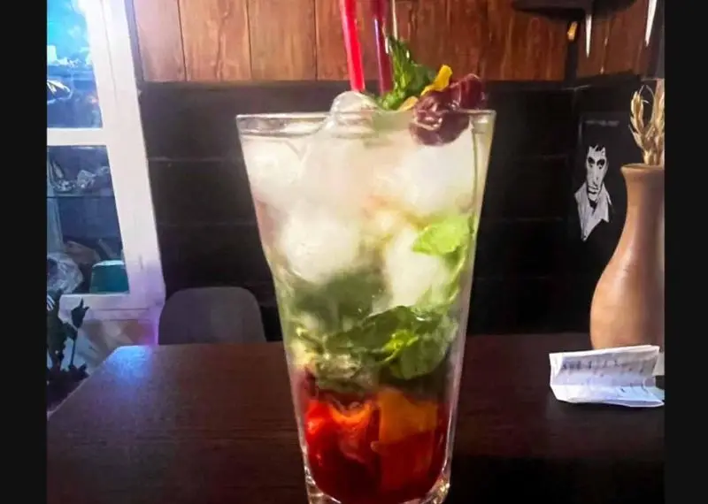 نوشیدنی خنک و میوه‌ای جذاب با تکه‌های میوه، نعنا و یخ در لیوان بلند روی میز کافه آگات با محیط دنج و دلنشین.
