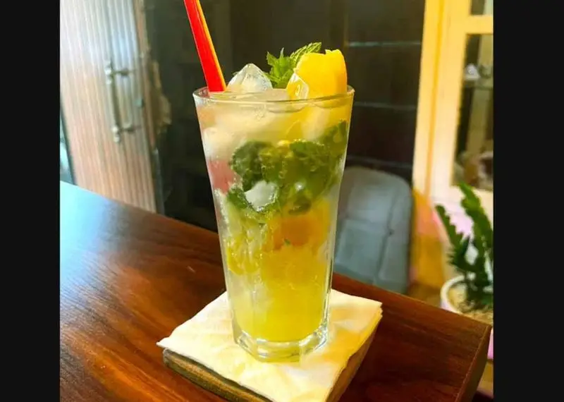 یک لیوان موهیتو میوه‌ای خنک و دلچسب با برگ‌های نعنا، تکه‌های میوه زرد و یخ در کافه آگات، نوشیدنی ایده‌آل برای آرامش.