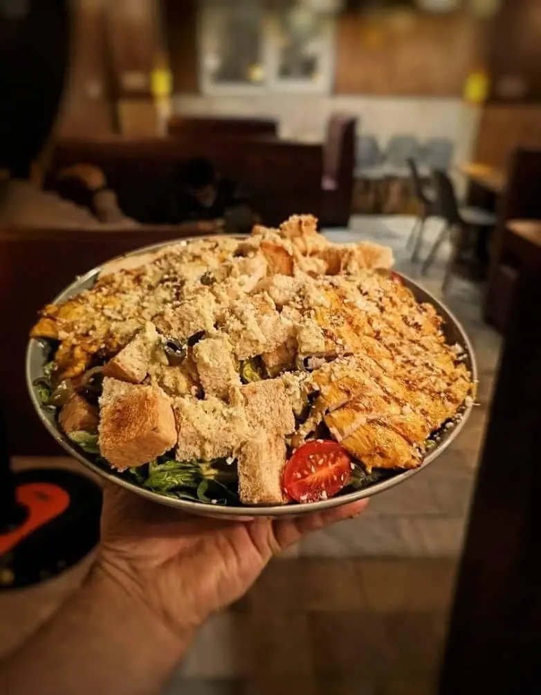 یک کاسه بزرگ سالاد سزار هیجان‌انگیز کافه آگات، پر از مرغ گریل، نان تست ترد و پنیر، آماده سرو در فضای دنج و زیبای کافه.