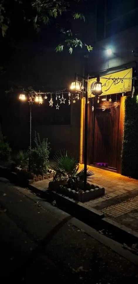 تصویری جذاب از ورودی کافه آگات در شب، با نورپردازی گرم و فانوس‌های آویزان به همراه تزئینات ستاره‌ای و ماه، فضایی دنج و رمانتیک.