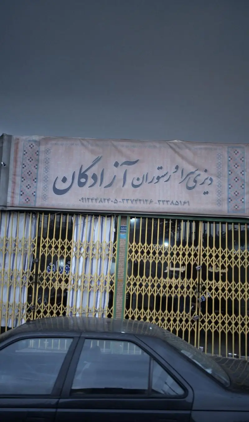 دیزی سرا و رستوران آزادگان، تابلوی کسب و کار با کرکره‌های زرد بسته و یک ماشین مشکی در نمای جلو. شماره تماس‌ها روی تابلو است.