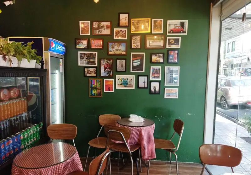 محیط داخلی دنج و دلنشین ساندویچ باماهاس با دیوار سبز گالری‌گونه، پر از قاب عکس‌های متنوع و میز و صندلی‌های چوبی شیک.