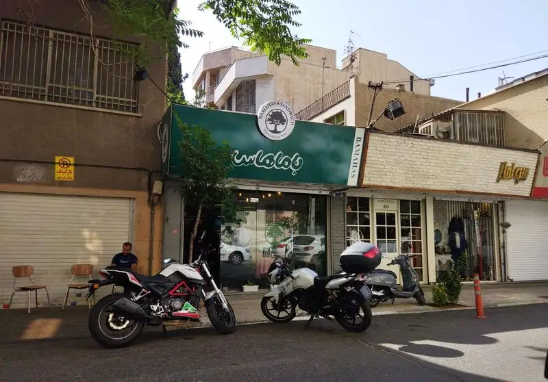 تصویری از نمای بیرونی ساندویچ باماهاس با تابلوی سبز رنگ و لوگوی درخت، همراه با چند موتورسیکلت در جلوی مغازه فست فود.