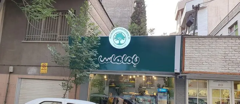 نمای بیرونی مغازه فست فود ساندویچ باماهاس در خیابان، با تابلوی سبز رنگ، لوگوی درخت و نوشته انگلیسی "Enjoy Life Your Way".