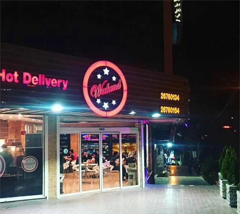 تصویری از رستوران فرنگی و فست فود ویکند در شب، با تابلوهای نئونی Hot Delivery، شماره‌های تماس و مشتریان داخل.