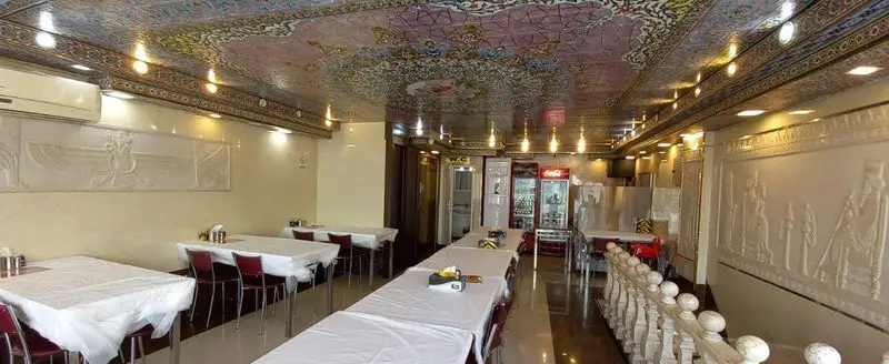 تصویری از داخل رستوران ایرانیان با دکوراسیون زیبای سنتی، میزهای غذاخوری مرتب، سقف نقش‌دار و حجاری‌های دیواری با نماد فرورهر.