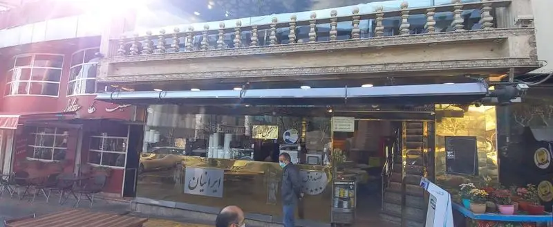 تصویر نمای بیرونی رستوران ایرانیان، با سردر و بالکن سنگی شیک، پیشخوان طلایی و تابلو "ایرانیان". مردی با ماسک در حال عبور از جلوی این رستوران محبوب.