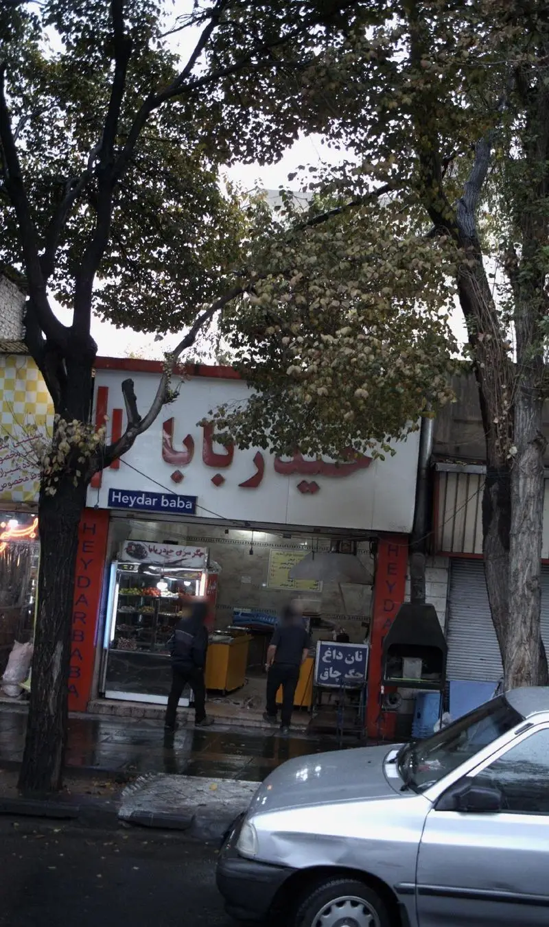 نمای بیرونی ویتامین سرای حیدر بابا، فروشگاه آبمیوه و جگرکی با تابلو Heydar Baba و نان داغ جگر چاق، درختان سبز در خیابان شلوغ