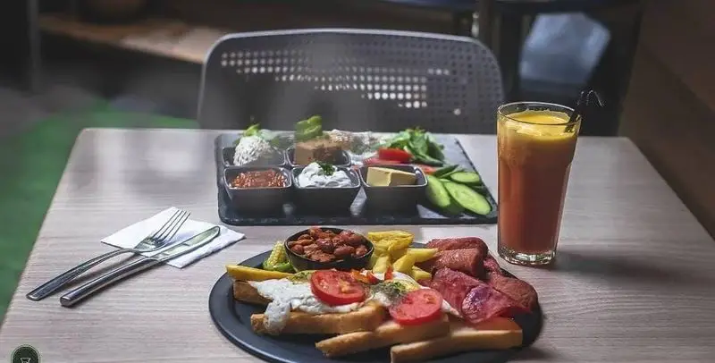 نمایی از یک میز پر از صبحانه کامل و متنوع در کافه رستوران آژمان با سوسیس، نان تست، آبمیوه، پنیر و مخلفات گوناگون.