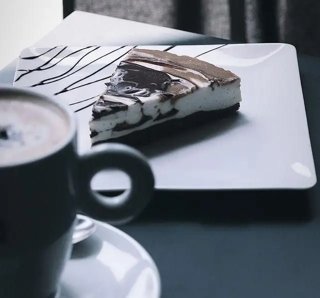 تجربه لذت بخش یک برش کیک شکلاتی و خامه ای با طرح مرمری و سس شکلات، همراه با فنجان قهوه، در محیط دلنشین کافه گالریا.