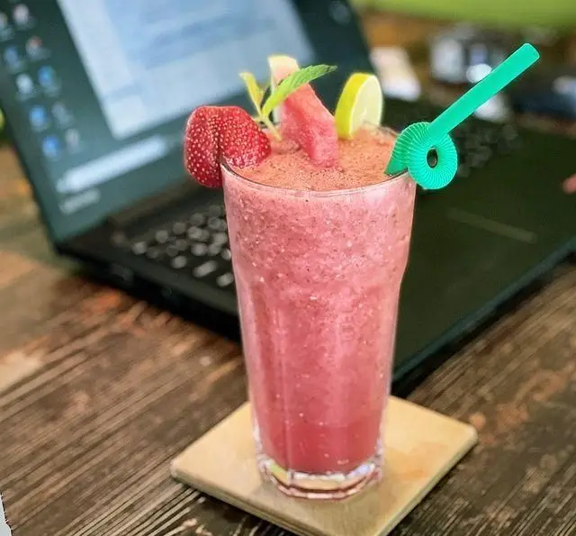 اسموتی توت فرنگی و هندوانه تازه در لیوانی بلند، روی میز چوبی کافه گالریا با لپ تاپ در پس زمینه، ایده‌آل برای کار.