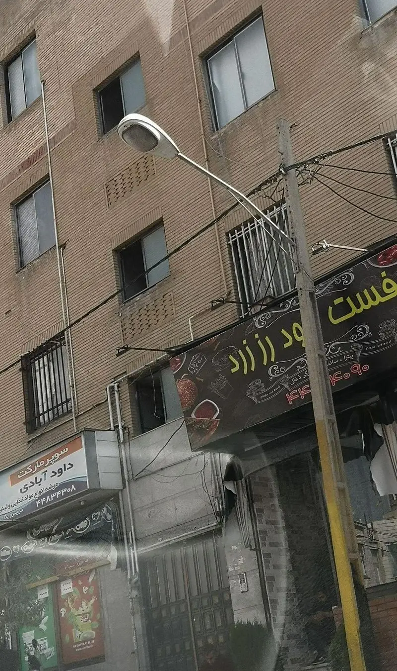 نمای شهری از فست فود پیتزا ساندویچ رز زرد، شامل تابلوی بزرگ مغازه، ساختمان‌های آجری و تیر برق در خیابان.