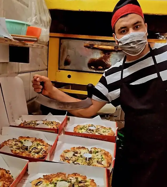 تصویری از آشپز "پیتزا ساندویچ رز زرد" با کلاه و ماسک، در حال برش پیتزاهای داغ با قارچ و فلفل در جعبه، کنار فر.