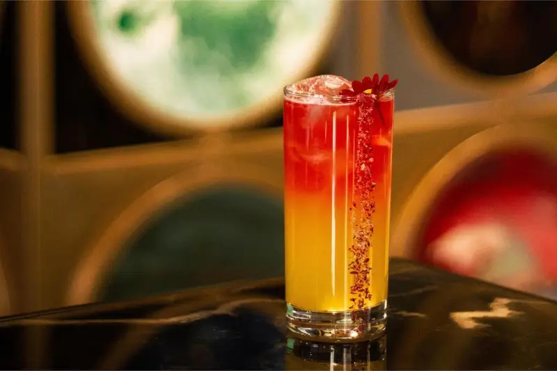 کوکتل جذاب و خوش‌رنگ با لایه‌های قرمز و نارنجی-زرد، تزئین شده با گل و ادویه، در فضای دلنشین کافه رستوران هوکاجی.