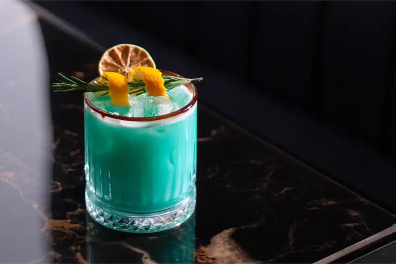 کوکتل آبی رنگ و خنک کافه رستوران هوکاجی با تزئین لیمو خشک، پوست پرتقال و رزماری، سرو شده روی میز مرمر قهوه‌ای. نوشیدنی دلنشین و خاص.