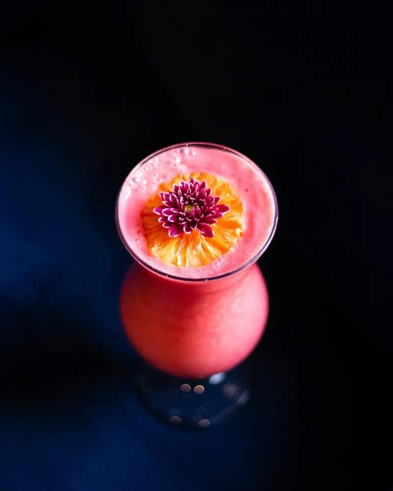 نمای بالا از کوکتل صورتی و خوشرنگ کافه رستوران هوکاجی، تزئین شده با گل ارغوانی و اسلایس پرتقال. منوی نوشیدنی‌های تازه و خنک.