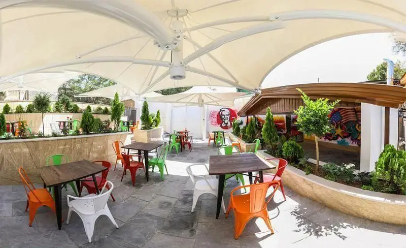 فضای باز فست فود چیتر چیکن با میز و صندلی‌های رنگارنگ زیر سایبان سفید، مناسب برای لذت بردن از غذای خوشمزه در محیطی دلنشین و سرسبز.