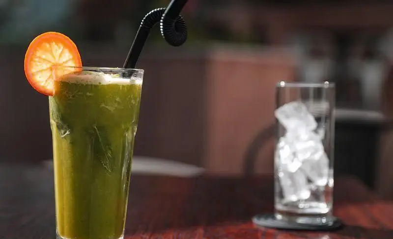 نوشیدنی سبز خنک و گوارا با برش پرتقال برای فست فود چیتر چیکن، همراه با لیوانی پر از یخ روی میز چوبی.