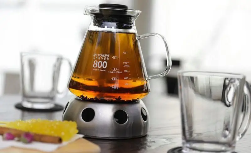 قوری چای شیشه‌ای مدرن روی وارمر به همراه نبات و دارچین. یک نوشیدنی گرم و دلچسب برای تکمیل وعده غذایی شما در فست فود چیتر چیکن.