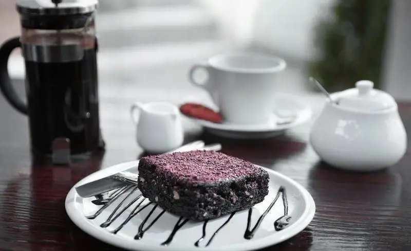 یک برش کیک شکلاتی تیره غنی با پودر تزیینی بنفش و سس شکلاتی، کنار قهوه تازه دم فرانسوی در کافه چیتر چیکن.