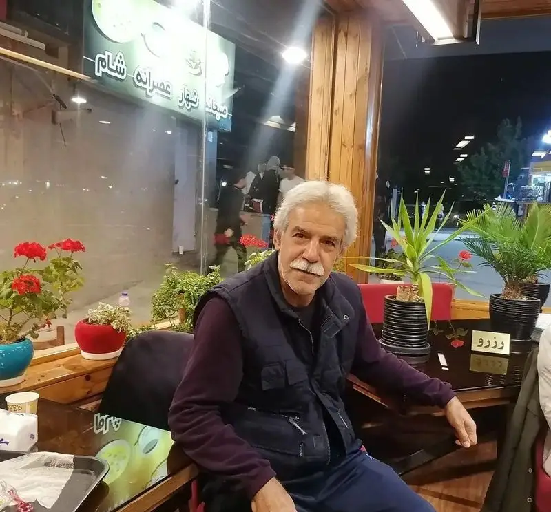 مردی با موهای خاکستری و سبیل در کافه رستوران استیج نشسته است. این تصویر فضای داخلی دنج و دلپذیر این کسب و کار را در شب نشان می‌دهد.