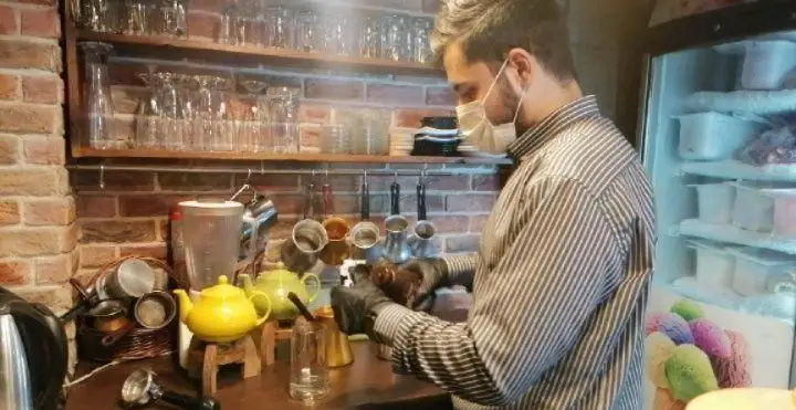 باریستای حرفه‌ای کافه رستوران استیج در حال تهیه قهوه‌ای خوش‌طعم با رعایت کامل پروتکل‌های بهداشتی، شامل ماسک و دستکش.