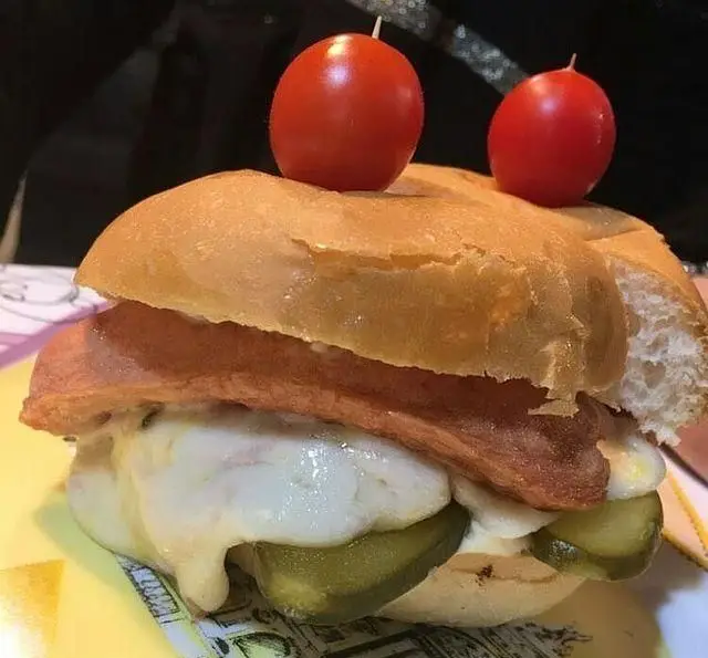 ساندویچ هات داگ داغ کافه رستوران استیج با پنیر آب شده و خیارشور، تزیین شده با شمع‌های فانتزی گوجه‌ای، انتخابی لذیذ و خلاقانه.