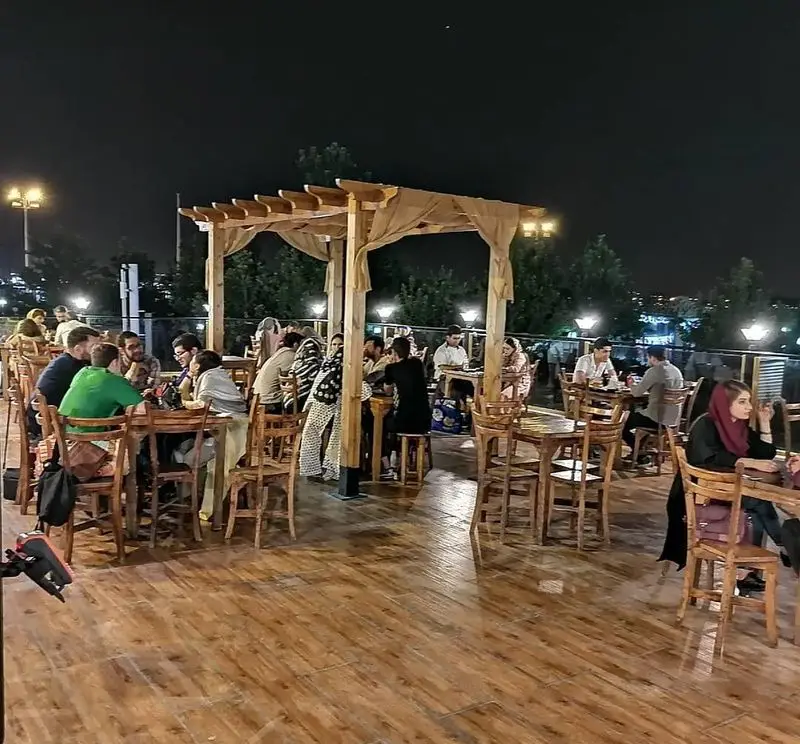 نمایی شبانه از کافه رستوران استیج؛ فضای باز پرجنب و جوش با میز و صندلی چوبی، مهمانان در حال گپ و گفتگو در محیطی دلنشین.