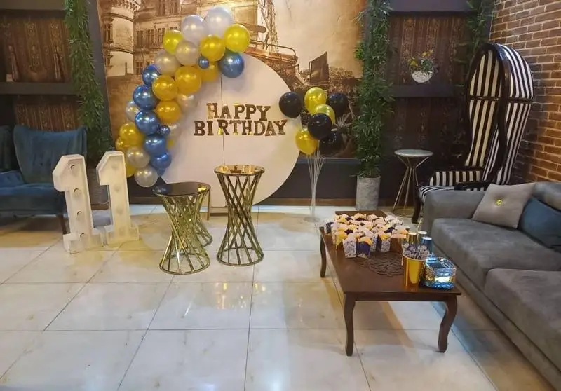 دکوراسیون شیک و جذاب جشن تولد 11 سالگی با بادکنک آرایی آبی، طلایی و سفید در کافه پناه، شامل میز دسر و مبلمان راحتی.
