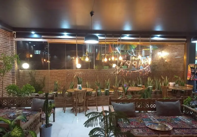 دکوراسیون شیک و تلفیقی کافه پناه: فضای گرم و دنج با مبلمان سنتی و مدرن، گیاهان آپارتمانی، دیوارهای آجری و نورپردازی خاص با چترهای آویز.