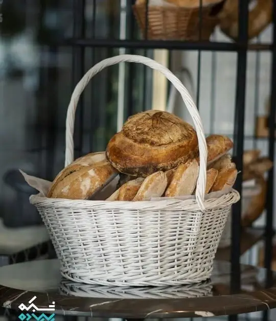 سبد حصیری سفید رنگ مملو از نان‌های تازه و خوش پخت کافه نانوایی پاتیس، شامل نان خمیر ترش، باگت و نان‌های کوچک. مناسب صبحانه و دورهمی.