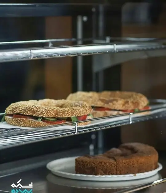 ساندویچ‌های سیمیت کنجدی تازه با سبزیجات و کالباس در ویترین کافه نانوایی پاتیس، همراه با کیک شکلاتی لذیذ برای صبحانه یا عصرانه.