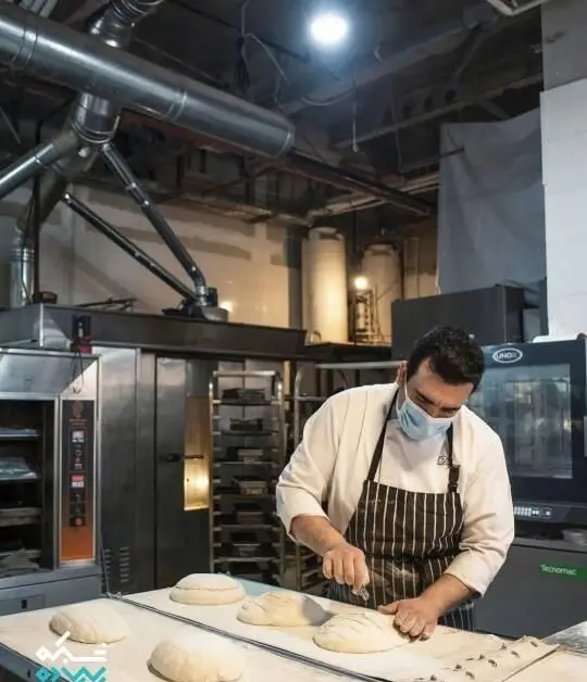 نانوای کافه نانوایی پاتیس در حال شکل‌دهی و برش نان‌های دست‌ساز برای پخت، با رعایت بهداشت کامل در آشپزخانه صنعتی